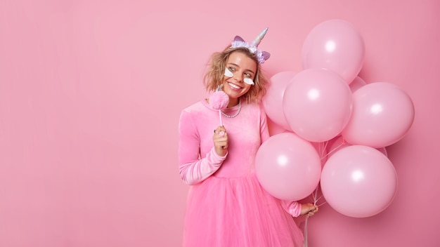 Positive verträumte junge Frau trägt festliche Kleidung, schaut weg, hat fröhlichen Ausdruck, trägt Schönheitspatches, trägt ein Einhorn-Stirnband, hält einen Haufen aufgeblasener Luftballons, die über rosafarbenem Hintergrund isoliert sind