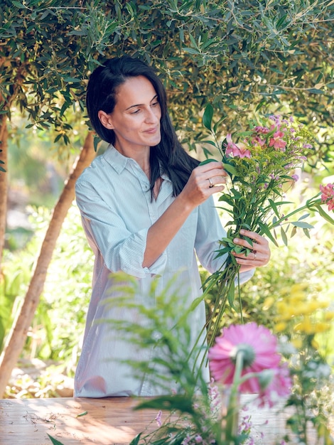 Positive weibliche Blumenhändlerin steht unter grünem Holz und arrangiert Blumen in Bündeln an einem sonnigen Sommertag im Garten