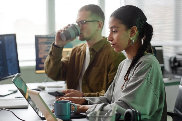 Positive weibliche Programmiererin benutzt Laptop zum Programmieren, während sie neben einer Kollegin im Büro arbeitet