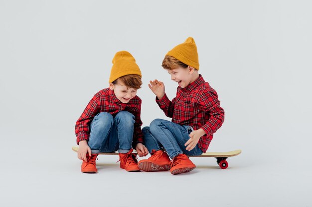 Positive Zwillinge in stylischem Outfit entspannen sich auf Skateboards auf weißem Hintergrund im Studio und lachen...