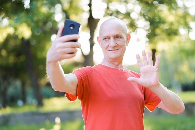 Positiver älterer Mann, der online kommuniziert und Hallo im Park zeigt.