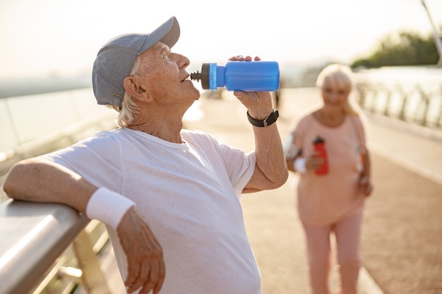 Positiver älterer Mann trinkt Wasser, das sich auf das Geländer stützt, während die Dame über die Fußgängerbrücke läuft