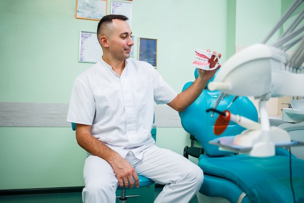 Positiver Arzt in weißer Uniform, der einen Scheinkiefer mit Zähnen zeigt, während er in der Zahnarztpraxis sitzt