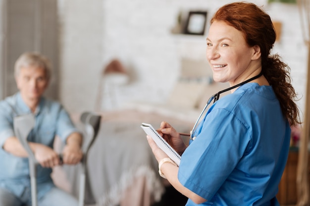 Positiver Behandlungsansatz. Begeisterte, kluge, erstaunliche Frau, die ihr Tagebuch verwendet, um die Symptome des Patienten aufzuschreiben, während sie einen Trost führt