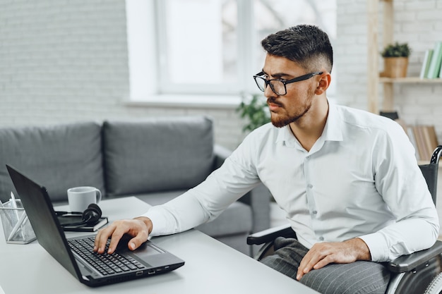 Positiver behinderter junger Mann im Rollstuhl, der im Büro mit Laptop arbeitet