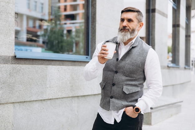 Positiver brutaler bärtiger grauhaariger älterer Mann, der Kaffee im städtischen Raum trinkt