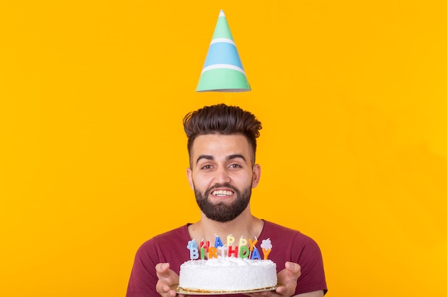 Positiver gutaussehender junger Hipster-Mann im burgunderroten T-Shirt, das Glückwunschkuchen mit der Aufschrift alles Gute zum Geburtstag hält, die auf einem gelben Hintergrund posiert. Konzept der Glückwünsche und Jubiläen.