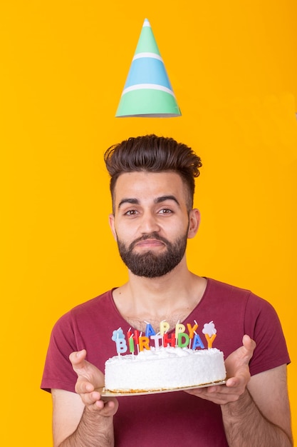 Positiver gutaussehender junger Hipster-Mann im burgunderroten T-Shirt, das Glückwunschkuchen mit der Aufschrift alles Gute zum Geburtstag hält, die auf einem gelben Hintergrund posiert. Konzept der Glückwünsche und Jubiläen.