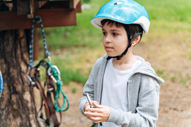 Positiver Junge in einem Schutzhelm steht in einem Seilpark und bereitet sich auf Unterhaltung vor. Das Kind verbringt aktiv Zeit