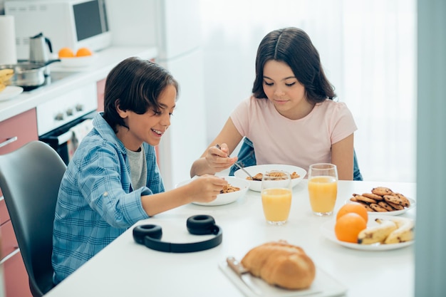 Positiver Junge und Mädchen sitzen am Küchentisch mit Schüsseln Cornflakes und Gläsern Orangensaft