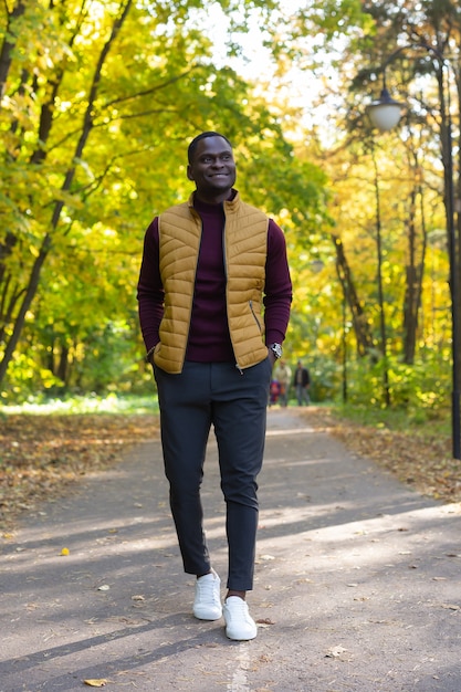 Positiver junger Afroamerikaner junger Mann in den stilvollen Kleidern geht im Spätsommerpark am sonnigen Tag.