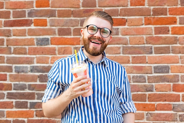 Positiver junger Hipster-Typ in Brille und mit Bart hält einen Cocktail mit einer Pfeife gegen eine Mauer. Das Konzept der Ferien und Sommerferien.