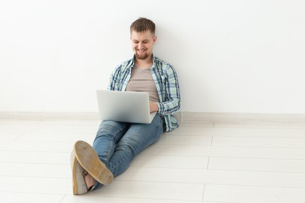 Positiver junger Mann in Freizeitkleidung, der im Internet auf der Suche nach neuen Wohnungen auf dem Boden in einem leeren Raum surft. Das Konzept, eine Wohnung über das Internet und einen Laptop zu finden. Exemplar