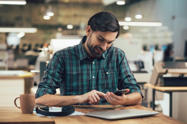 Positiver junger Mann in kariertem Hemd, der mit Kaffee und Kopfhörern am Bürotisch sitzt und lächelt, während er auf den Bildschirm eines Gadgets schaut