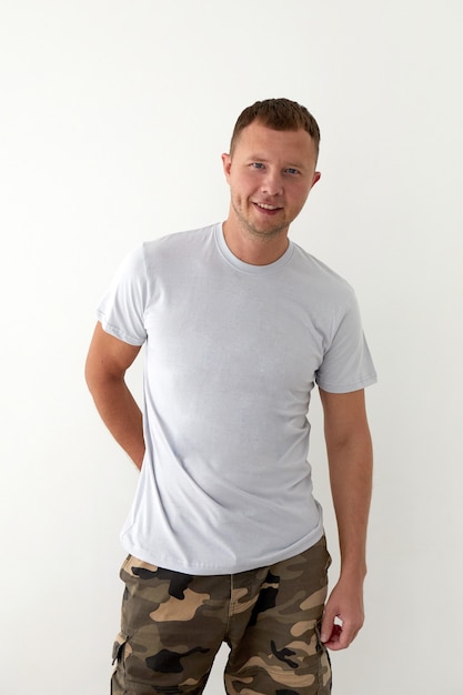 Positiver junger Mann in khakifarbener Militärhosenhand hinter und grauem T-Shirt, das vor weißem Hintergrund steht und in die Kamera schaut