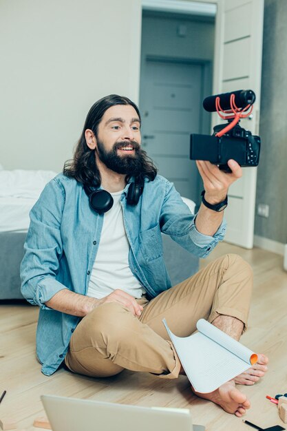 Positiver junger Mann mit Notizbuch und einem Laptop, der zur Kamera in seiner Hand lächelt