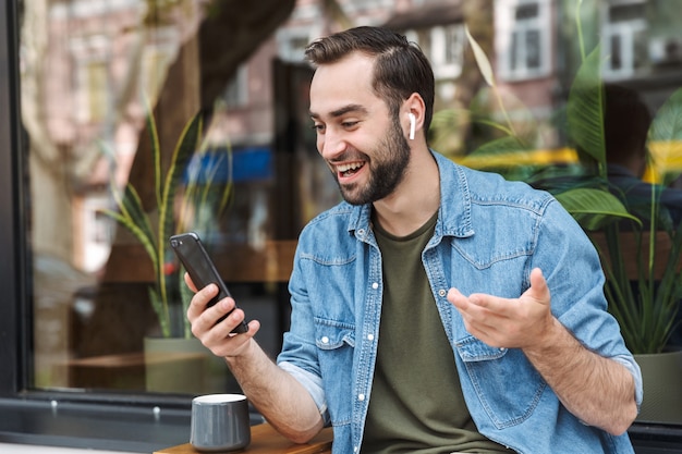 positiver junger Mann mit Ohrstöpseln, der auf dem Smartphone hält und spricht, während er im Freien im Stadtcafé sitzt