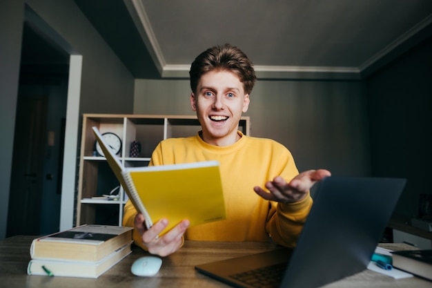 Positiver junger Student, der zu Hause an einem Schreibtisch sitzt, mit einem Notizbuch in den Händen und einem Laptop, der mit einem überraschten Gesicht in die Kamera schaut Emotionaler Student studiert zu Hause