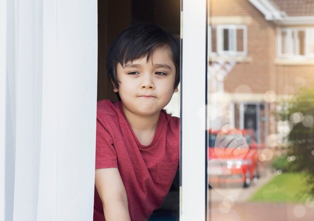 Positiver Kinderjunge sitzt am Fenster und schreibt auf Glas und schaut mit lächelndem Gesicht hinaus