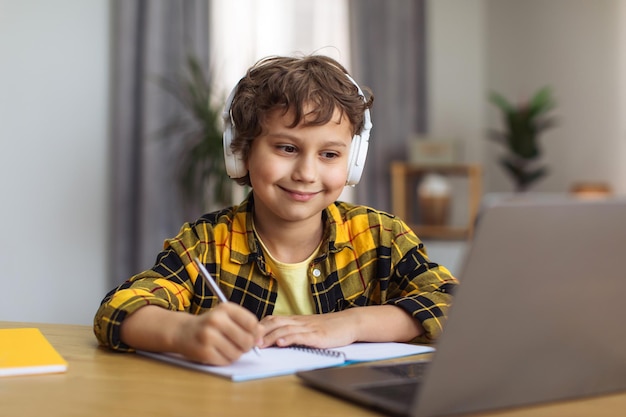 Positiver kleiner Junge, der Übungen aufschreibt, die Online-Lektionen auf dem Laptop zu Hause beobachten, Nahaufnahme, Porträt