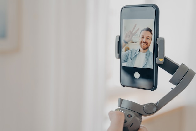 Positiver Mann-Blogger, der einen Gimbal-Stabilisator mit Smartphone hält und versucht, Selfie-Bilder und Live-Videos zu machen, während er Zeit zu Hause im Wohnzimmer verbringt. Vlog- und Video-Blogging-Konzept