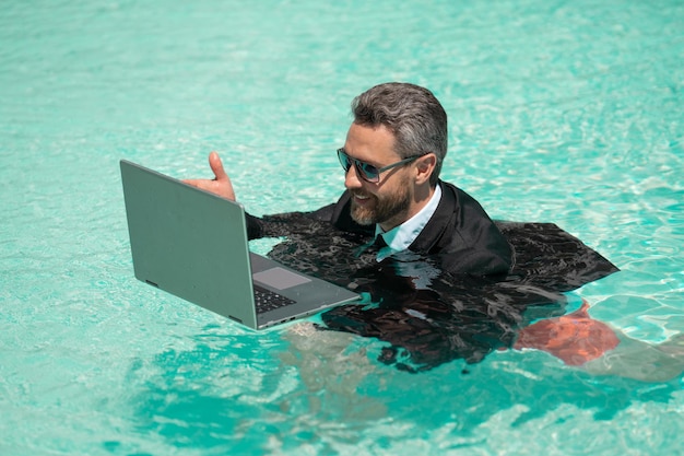 Positiver Mann hat am Wochenende einen geschäftlichen Videoanruf, um sich am Pool zu erfrischen. Mann hat am Wochenende einen geschäftlichen Videoanruf mit einem Laptop. Foto eines Mannes, der am Wochenende einen geschäftlichen Videoanruf hat. Mann hat am Wochenende einen geschäftlichen Videoanruf