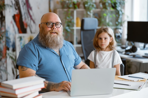 Positiver Mann mit Brille hilft kleiner Tochter, Hausaufgaben am Laptop am Tisch zu machen