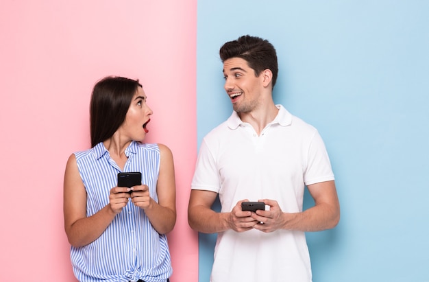 positiver Mann und Frau in der Freizeitkleidung stehend und halten Smartphones, lokalisiert über bunte Wand