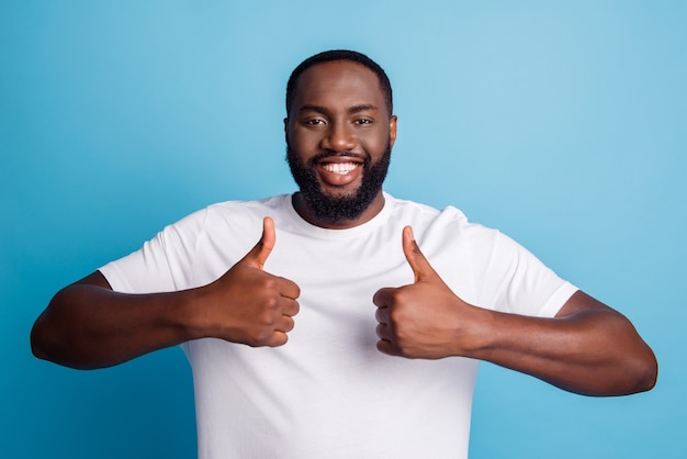 Positiver netter froher Afro-Mann hebt zwei Daumen hoch glänzendes weißes Lächeln posiert auf blauem Hintergrund