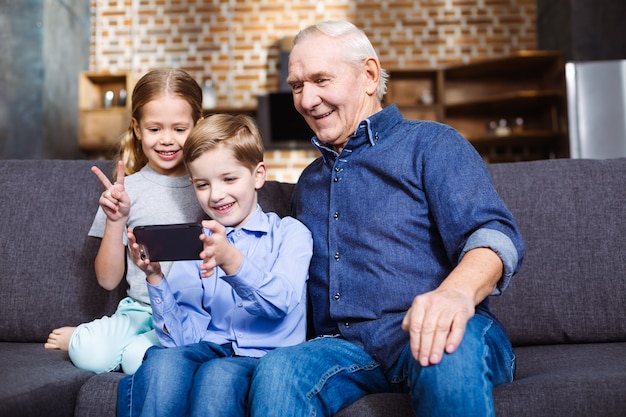 Positiver pensionierter mann, der auf der couch sitzt, während er mit seinen enkelkindern selfies macht