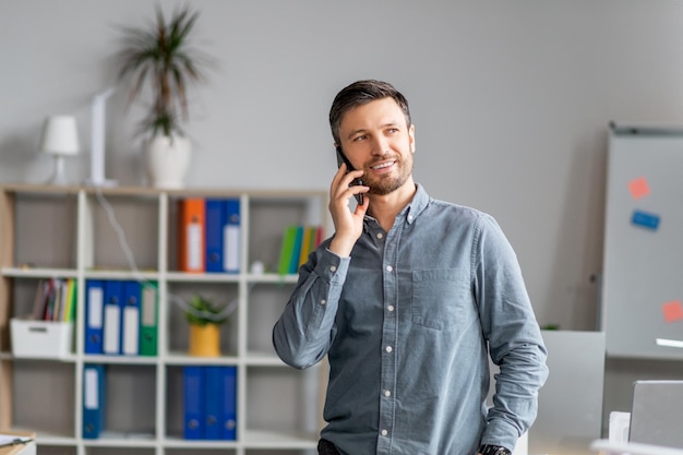 Positiver reifer Mann im Büro, der am Handy in der Nähe des Arbeitsplatzes spricht und sich mit dem Kunden unterhält