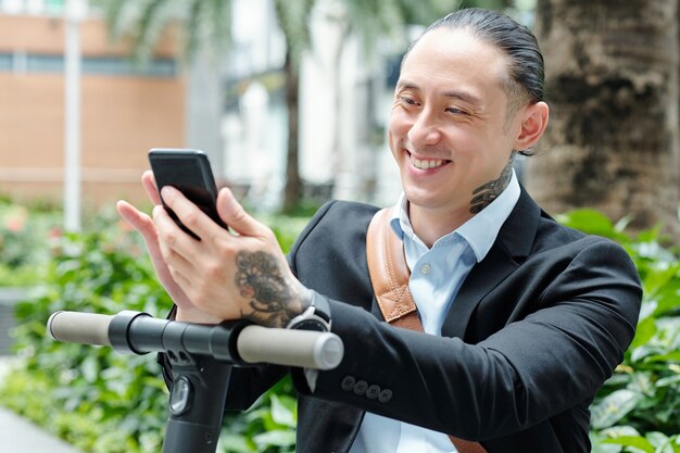 Positiver stilvoller Geschäftsmann der gemischten Rasse, der Nachrichten im Smartphone prüft, nachdem er auf Roller gefahren wird