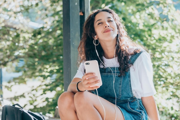 Positives afrikanisches glückliches junges Studentenmädchen, das Denim-Overalls trägt, das auf dem Campus sitzt, afrikanische junge Frau hört Musik mit Kopfhörern, die drahtloses Entspannen des Mobiltelefons verwendet, Kopienraum