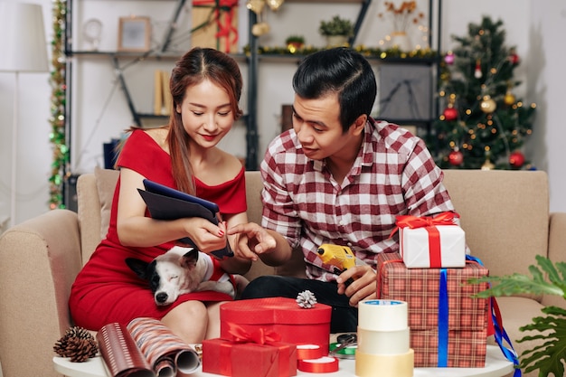 Positives asiatisches Paar mit kleinem Hund nach dem Tutorial auf dem Tablet-Computer beim Dekorieren von Weihnachtsgeschenken und Geschenken für Familienmitglieder