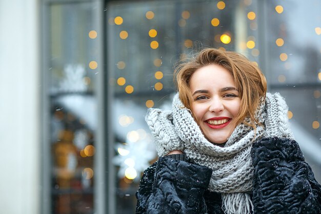 Positives brünettes Mädchen mit roten Lippen trägt Mantel und Schal, das bei Schneefall in der Stadt spazieren geht. Freiraum