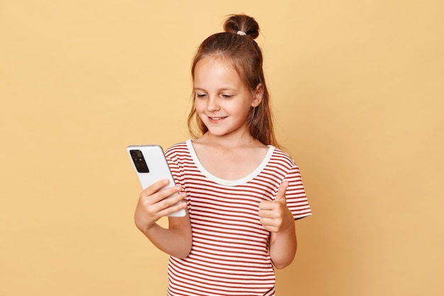 Positives, glücklich lächelndes kleines Mädchen mit braunen Haaren, das ein gestreiftes T-Shirt trägt und isoliert vor beigem Hintergrund steht und ein Mobiltelefon benutzt, das wie eine Geste zeigt und Internetinhalte genießt