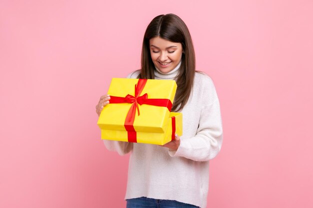 Positives, glückliches, dunkelhaariges Mädchen, das in eine verpackte Geschenkbox schaut, offenes Geschenk, positiv ausdrückt, einen weißen, lässigen Pullover trägt. Studioaufnahme im Innenbereich isoliert auf rosa Hintergrund.