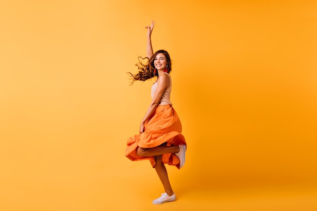 Positives, gutaussehendes Mädchen, lustiges Tanzen auf gelbem Hintergrund Prächtige weiße Frau in langem orangefarbenem Rock, die mit aufrichtigem Lächeln springt