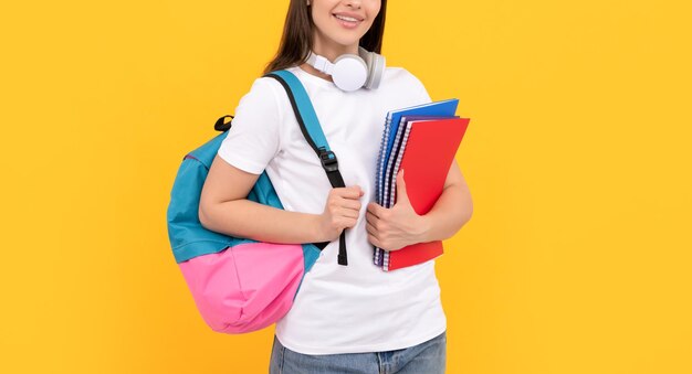 Positives junges Mädchen mit Rucksack und Kopfhörern halten Arbeitsbuch auf gelber Hintergrundschule