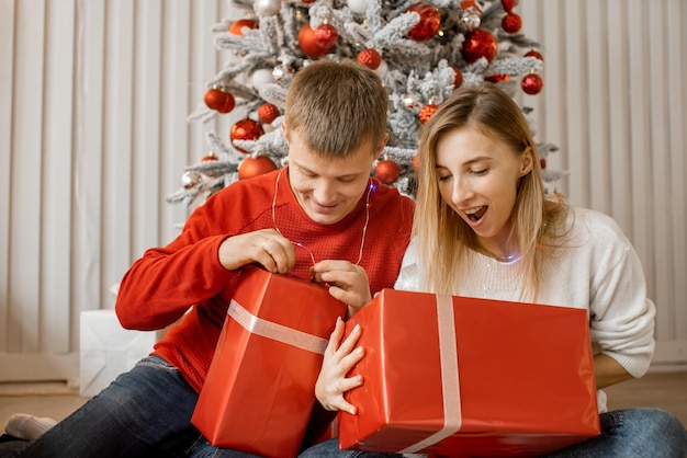 Positives junges Paar, das neben dem Weihnachtsbaum auf dem Boden sitzt und voller Aufregung Geschenke auspackt