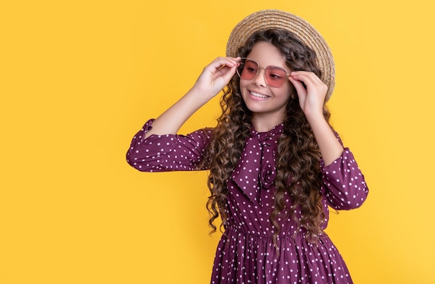 Positives Kind mit Strohhut und Sonnenbrille mit langen brünetten lockigen Haaren auf gelbem Hintergrund