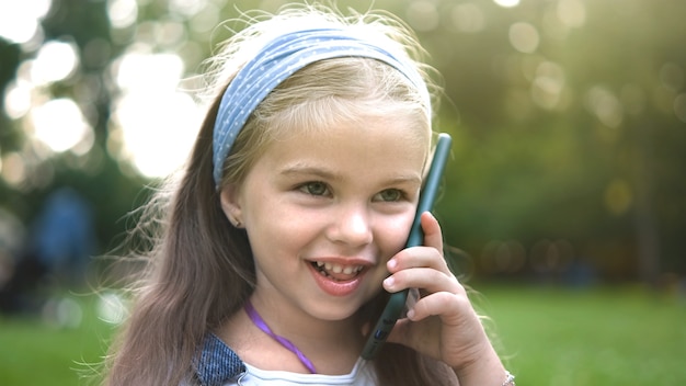 Positives kleines Mädchen mit Gespräch auf ihrem Handy im Sommerpark. Junges weibliches Kind, das auf Sellphone spricht.