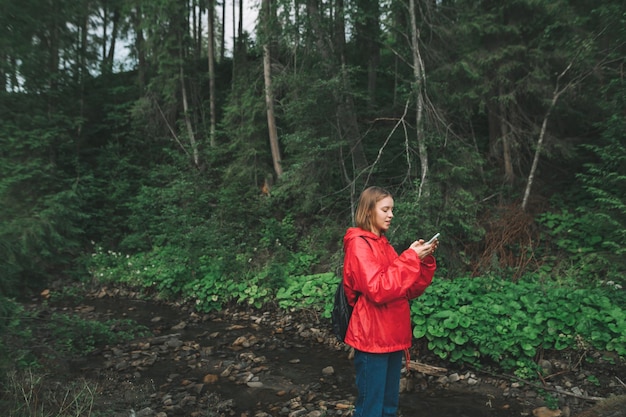 Positives Mädchen in einem roten Regenmantel steht im Wald in der Nähe eines Bergbachs und benutzt ein Smartphone mit einem Lächeln im Gesicht Frauentourismus nutzt das Internet bei einer Wanderung im Schoß der Karpaten