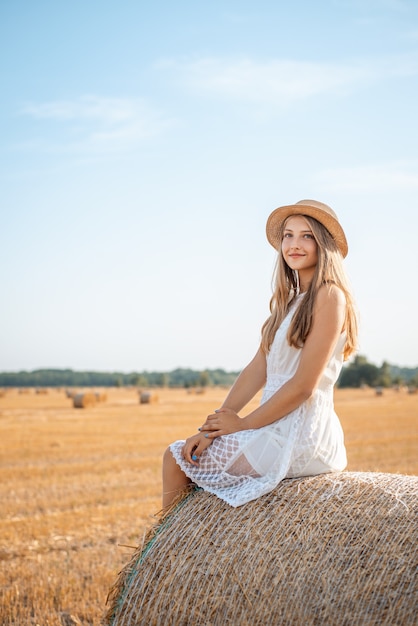 Positives Mädchen in einem weißen Kleid und einem Strohhut, die auf einem Strohballen auf dem landwirtschaftlichen Feld sitzen