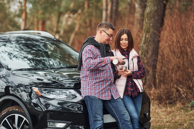 Positives reifes Paar, das in der Nähe ihres Autos mit warmem Getränk in den Händen steht Im Freien im Wald