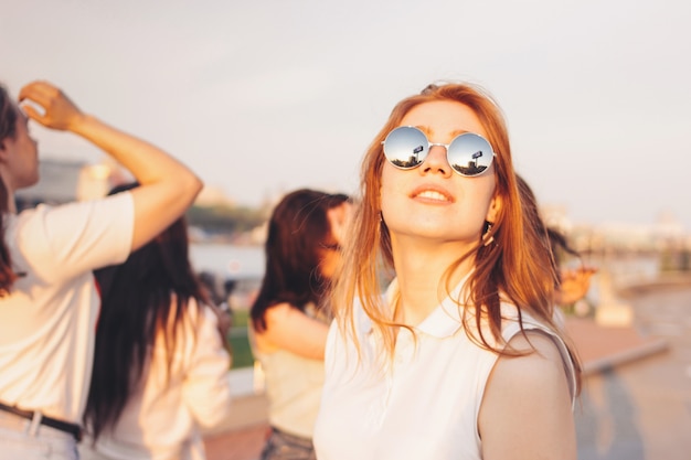 Positives schönes glückliches rotes behaartes Mädchen in der Spiegelsonnenbrille mit Freunden auf Hintergrund des blauen Himmels