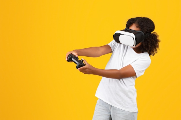Positives schwarzes Teenagermädchen in weißem T-Shirt, VR-Brille und Joystick genießt Freizeit beim Spielen