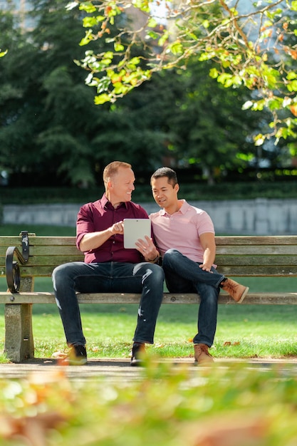 Positives schwules Paar, zwei Männer, die draußen auf einer Bank sitzen und ein digitales Tablet beim Online-Shopping halten