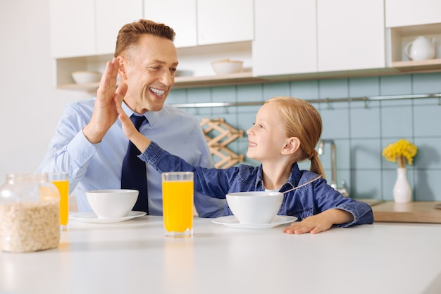 Positives süßes nettes Mädchen, das ihren Vater ansieht und ihm High Five gibt, während er mit ihm frühstückt