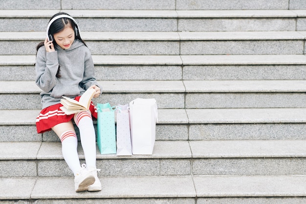 Positives Teenager-Mädchen mit Kopfhörern, das auf Stufen sitzt und nach dem Unterricht ein interessantes Buch liest?